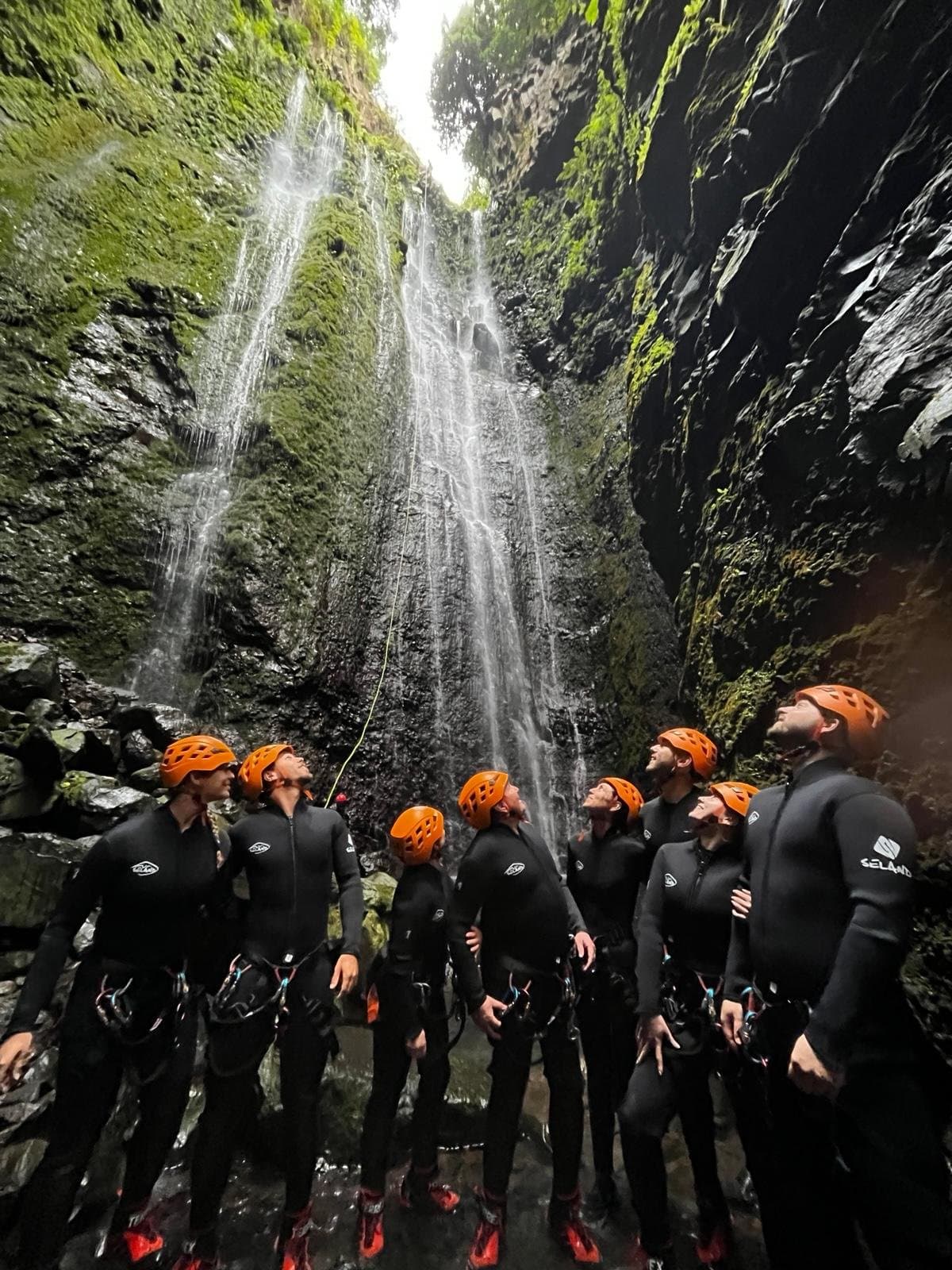 A group in wetsuits and orange helmets looking up at the big waterfall in Ribeira Funda