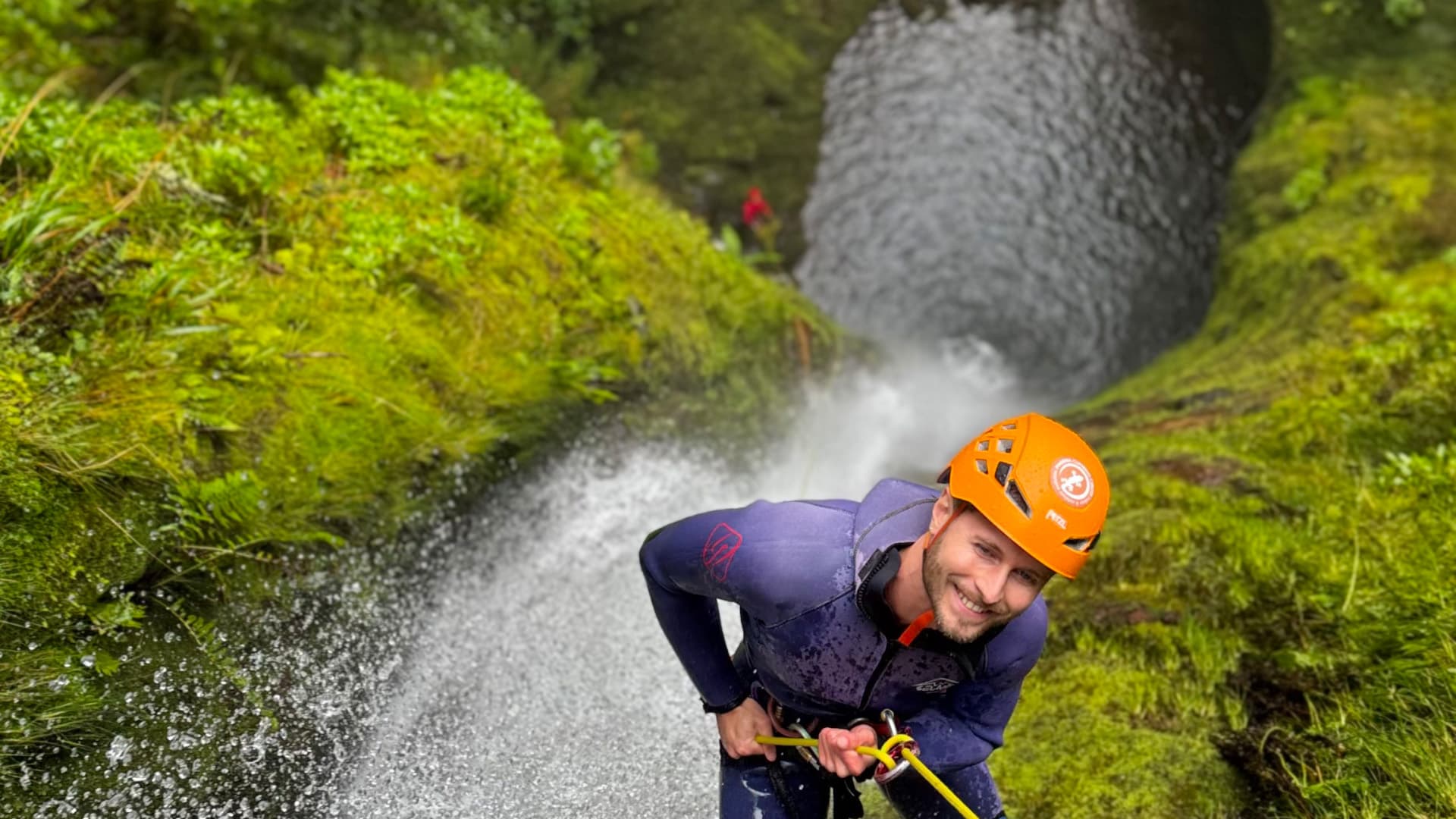 A smiling canyoner abseiling beside a waterfall in a moss-covered Madeira canyon